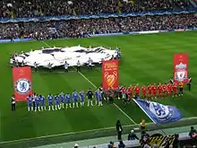 The Chelsea (blue) and Liverpool (red) teams line up side-by-side in front of banners bearing their club crests and the Champions League final logo. In the background, a flag bearing the Champions League logo is waved over the centre circle, while the main stand is further back, full of spectators. In the foreground, Chelsea fans wave blue flags bearing their club crest.