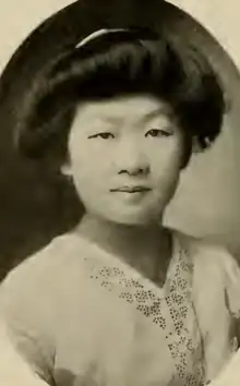 A young Chinese woman, dark hair dressed in a bouffant updo, wearing a v-necked white blouse with embroidered trim