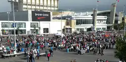 Several thousand people stand in lineups while volunteers serve food. A large shopping mall stands in the background.