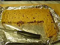 A foil-lined tray with a solid light brown sheet with dark brown spots. A section has been cut away and most of a knife is lying in that portion.