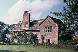A stone house seen from a slight angle, with a gabled wing to the right and a tall chimney on the roof to the left