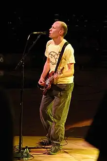 Chris Ballew wearing a yellow print t-shirt, dark pants, and sneakers, standing onstage playing guitar and singing into a microphone