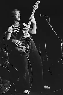 Brown is shown in a full-length, black-and-white shot. He is leaning backwards and towards his right, with the bass guitar held at a steep angle. A microphone is in shot in front of him.
