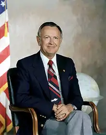 Formal portrait of a man in a jacket and tie seated in a chair in front of a U.S. flag.