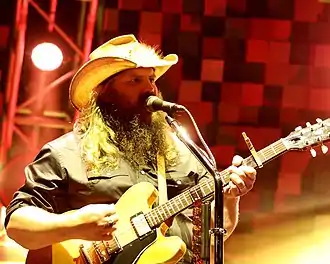 A man with long hair and a beard, wearing a cowboy hat, singing into a microphone and playing a guitar