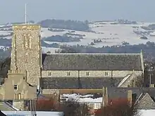 Side view of the upper section and tower of a long, flint-built church with snow-covered hills in the background and various rooftops in the foreground. A tall tower rises on the left.