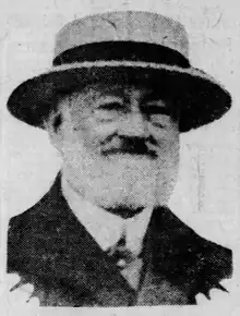 Newspaper photograph of a smiling elderly man with a white beard, wearing a suit and a hat.