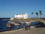 Several small boats float onshore. A white church with a red roof stands several metres away up a sandy path.