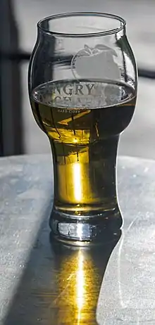A drinking glass with the Angry Orchard name and logo on it, bulbous at the top, half-filled with clear dark yellow liquid through which the sun shines, reflected on the metal surface in front of it
