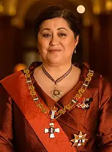 A smiling woman wearing a Māori feather cloak posing in front of a graphic of the New Zealand coat of arms