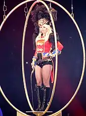 Image of a blond woman. She is standing with a red feathered jacket, carrying a whip around her neck and singing in a wireless microphone. Several people surround her, all wearing S&M outfits.