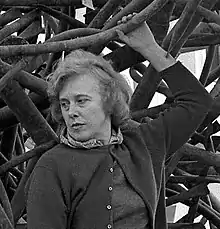 Claire Falkenstein, standing on one of her sculptures, her left arm raised above her head holding part of the sculpture