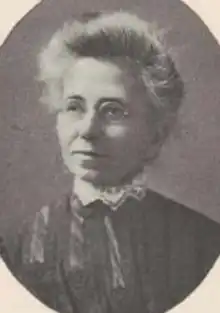 An older white woman, grey hair in a bouffant updo, wearing glasses and a high lace collar with a dark garment