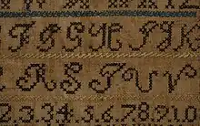 Close-up of an alphabet embroidery sampler made with blue, green, black, and yellow thread. The rows of letters are each separated by a line of herringbone stitches.