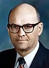 Color photo of a bald man wearing glasses and a suit with a striped tie