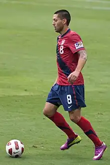 A man wearing a red shirt and blue short pants readies to kick a soccer ball