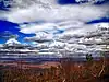 View from a high vantage point of a mountainous landscape beneath a party cloudy sky, there are twisted small trees in the foreground