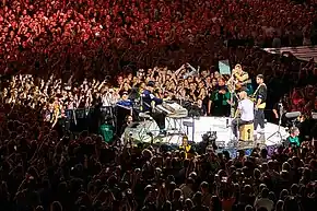 Four men perform on a small stage as a large crowd is gathered around them