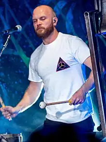 A bald, bearded man wearing a white shirt plays the timpani while a church bell is beside him