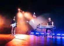 A woman performs with a microphone and guitar while a man plays the piano and sing with her