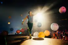 A man walks on the stage as giant colourful balls bouce in the background