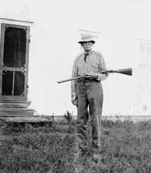 picture of a man with a moustache, standing, holding a rifle and wearing a hat