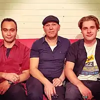 Three men are seated on a red-covered couch. The man at left has short, black hair and stares straight ahead with a slight smile. He has a red shirt over the top of a black tee-shirt and dark pants. The second man's head is slightly tilted to his right and he wears a black beret. He also stares straight ahead, he has a light stubble to his chin. He wears a dark blue shirt over a white tee-shirt and dark blue pants. The third man tilts his head further and sports longer, brown hair. He wears a dark blue, unbuttoned shirt over a tee-shirt with some lettering obscured, which advertises "Kiama Jazz & Blues Festival".