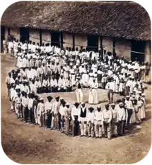 Photograph showing a group of people dressed in white, who have gathered in front of a tile-roofed farm building and observe another large group which has formed a large circle surrounding 5 men straddling large drums, a woman and 2 other men.