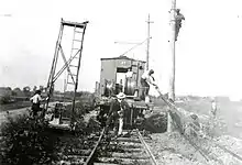 This black and white photograph shows construction workers raising power lines next to the railroad tracks of the Toledo, Port Clinton, Lakeside Railroad tracks in a rural area. The workers are using a railroad car as their vehicle to carry supplies and themselves down the line. It was taken in approximately 1920.