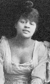 A young African-American woman with her hair in a top bun; her dress is light-colored with a low square neck.