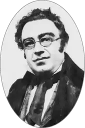 Round-faced white man, clean shaven with unruly dark hair and round spectacles