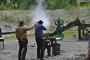 A Cowboy action shooter firing a lever action rifle at steel targets. The Range Officer to the left is holding a shooting timer to measure the time.
