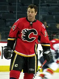 A man with short, brown hair looks to his right.  He is wearing a red uniform with black pants and a black stylized C logo on his chest.