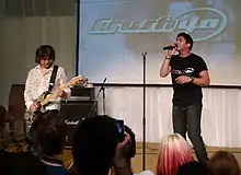A Japanese man (left) plays a guitar while an American man (right) sings in front of a crowd.