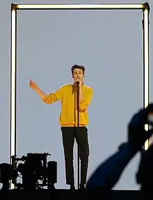Albert Černý wearing black pants and yellow sweater, standing in front of microphone, holding right arm out, looking at camera