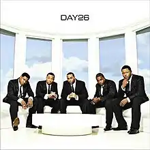 The cover shows the group dressed in black suits with neckties, sitting on a white couch near a table. Behind the group is a blue sky with clouds.