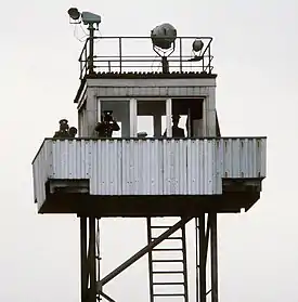 A metal observation tower manned by three GDR guards. Some watchtowers were semi-portable and could be moved to new sectors when needed.[citation needed]