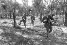 A number of armed soldiers moving in a tactical formation move through the trees towards some barbed wire.