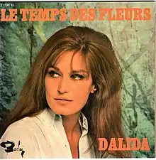 Dalida posing in studio in front of orange background, looking aside she wears 1960s style shirt and her signature minimalistic makeup around eyes.
