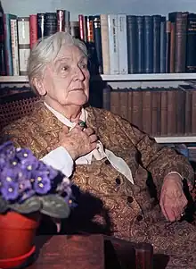 old white woman, seated in front of a well-filled bookcase