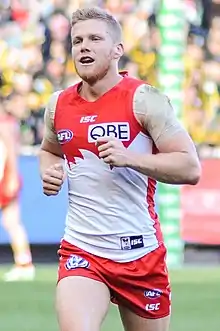 A male athlete with light hair wearing a sleeveless guernsey and shorts jogs on the grass surface of the playing arena.