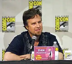  A Caucasian man in his forties, seated at a conference, with a microphone in front of him. He has a pleasant square face, deep-set eyes, dark hair and a brown beard with clean-shaved cheeks and upper-lip. He is casually dressed, relaxed and smiling. Square signs are posted on the wall behind him, bearing the name COMIC-CON in big bright yellow letters around a drawn eye and eyebrow.