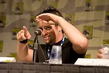 Man seated in front of microphone, shading his eyes and pointing to audience member