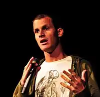 A dark haired Caucasian man wearing a white shirt, and green jacket while performing a stand-up comedy routine