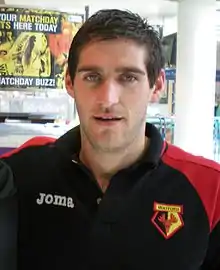 The head and upper torso of a young man, wearing a black and red top. The logo on his top is coloured red, black and yellow, and reads "WATFORD"