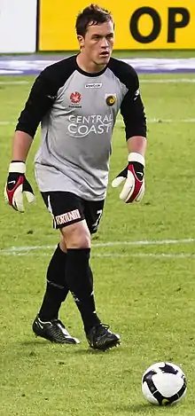 A man with dark hair, wearing a grey shirt, black shorts and goalkeeping gloves approaching a football