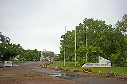 A large sign marking a pathway to Darwin High School, which is mostly covered in trees. There are three bare flagpoles near the sign.
