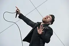 A color photograph of American musician Davey Havok performing live in 2014, dressed in all-black and holding a wired microphone.