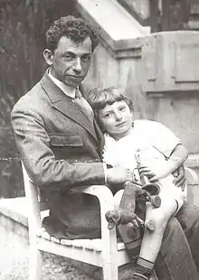 A man sitting on a chair with a small boy on his lap, the boy is hold a small toy dog, both are looking towards the viewer