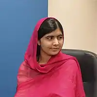 Malala seated in a chair facing away from the camera wearing a red head covering that drapes over her shoulders.